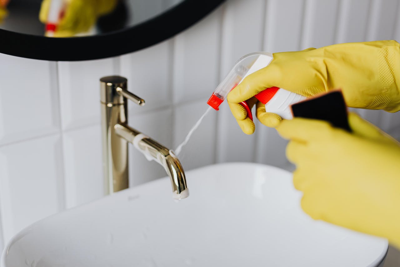 services-03 From above anonymous housekeeper in yellow rubber gloves spraying cleaner to chrome water faucet in bathroom