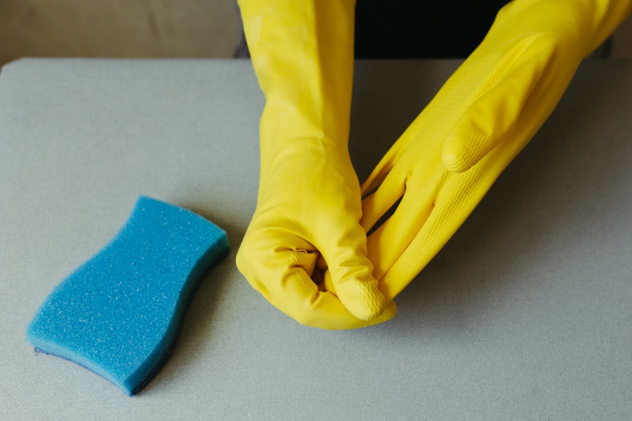 hero-img Close-up of hands wearing yellow gloves next to blue sponge on gray surface.
